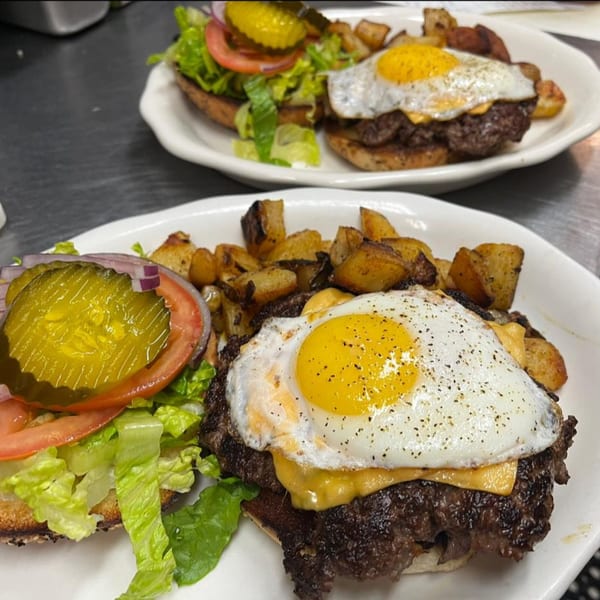 Two breakfast burgers with fried eggs, cheese, lettuce, tomato, pickles, and a side of home fries.
