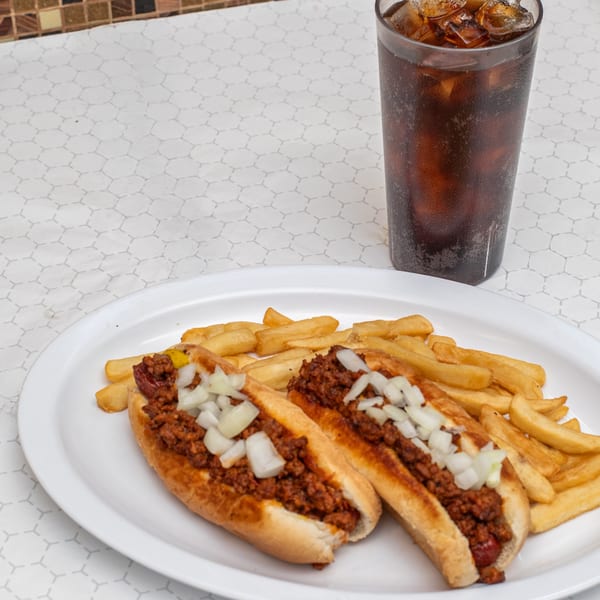 Two chili dogs with diced onions, a side of fries, and a glass of soda on a white plate.